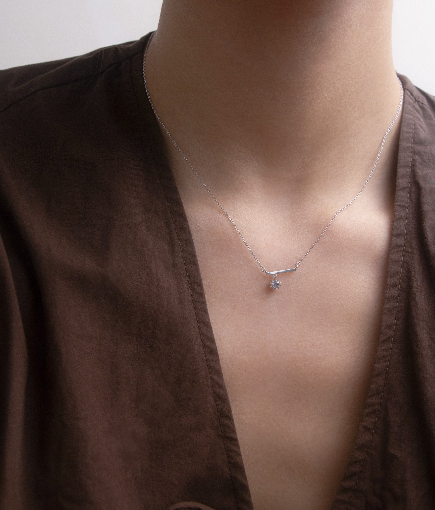 Close up front view of a silver necklace with a zirconia pendant worn by a person in a brown garment
