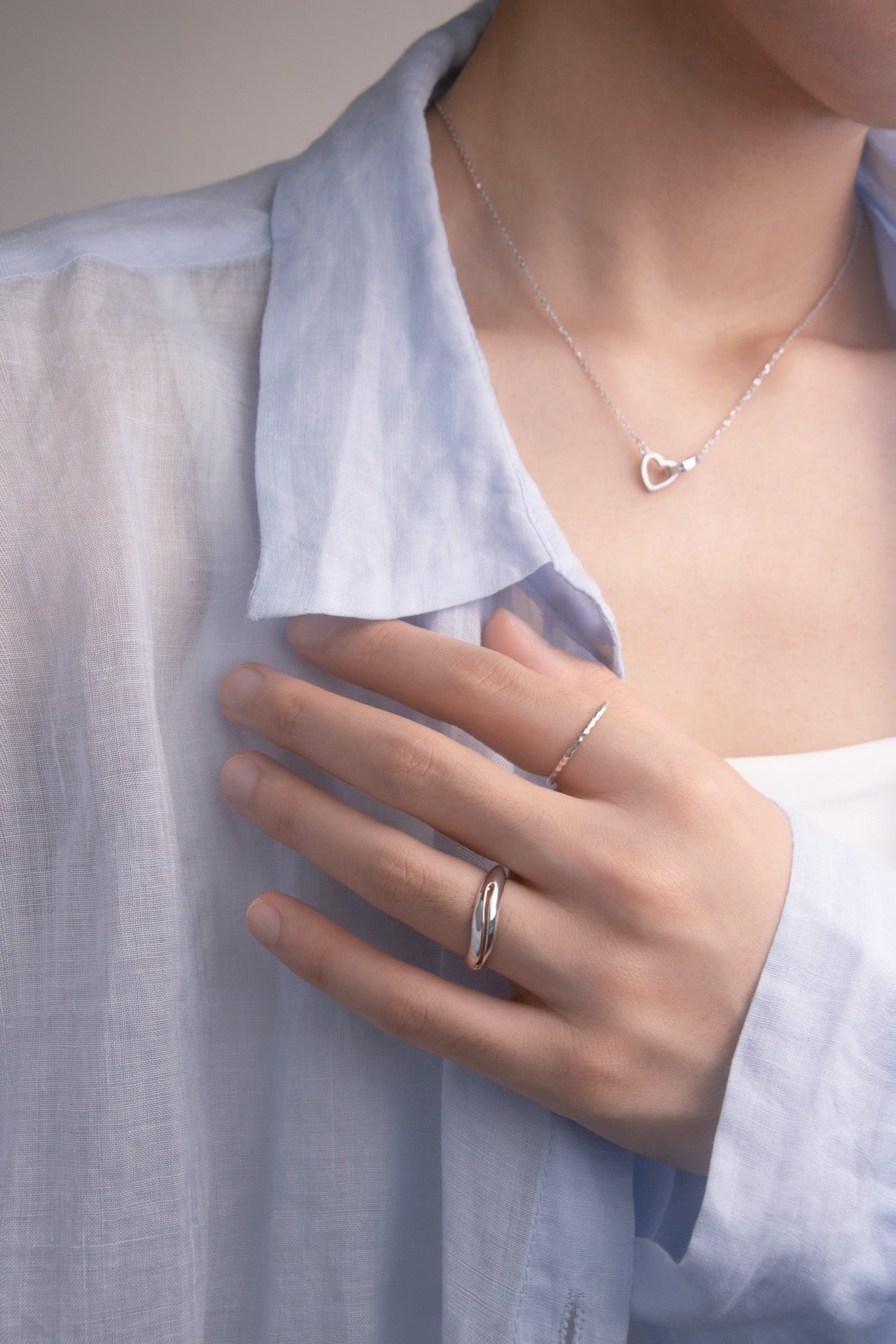 Close-up front view of a person wearing a silver necklace with a heart pendant and a silver ring on a light blue shirt.