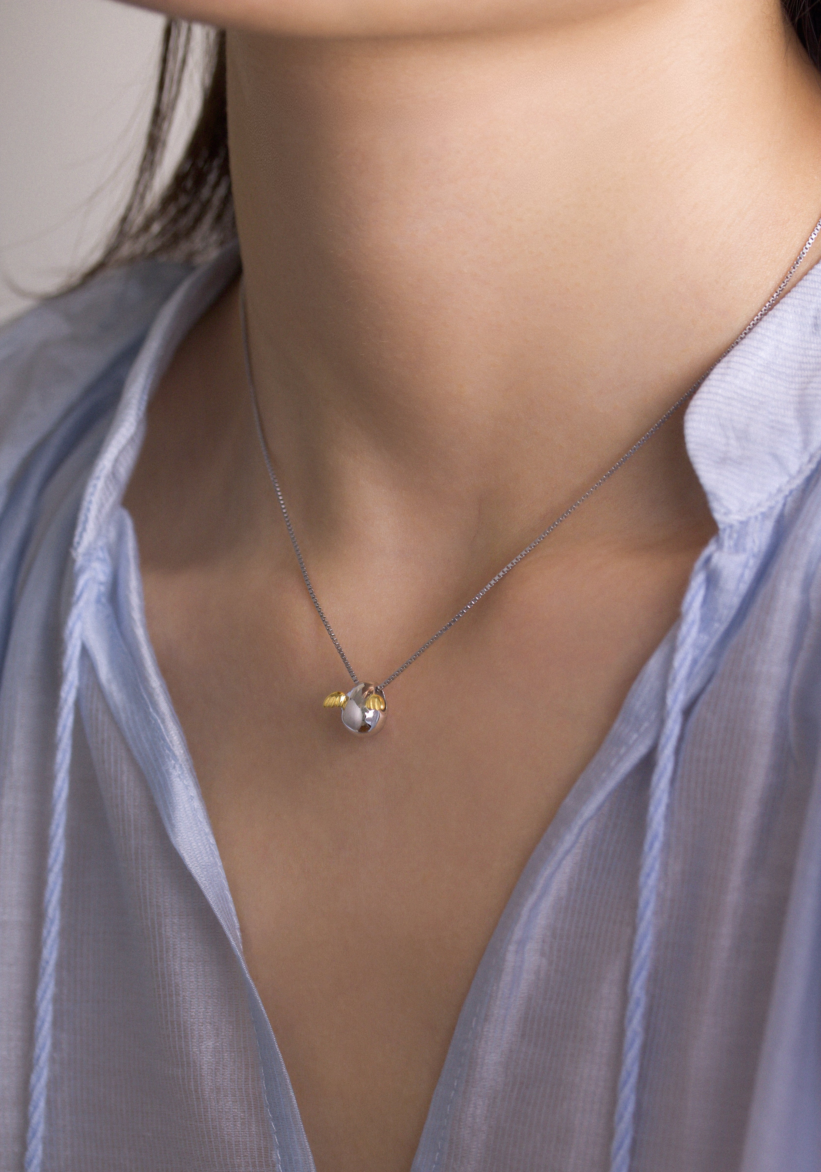 Fine jewellery, silver jewellery, hypoallergenic, tarnish-free, side view of a silver Necklace with a pendant with golden wings worn by a person wearing a light blue shirt on the neck.