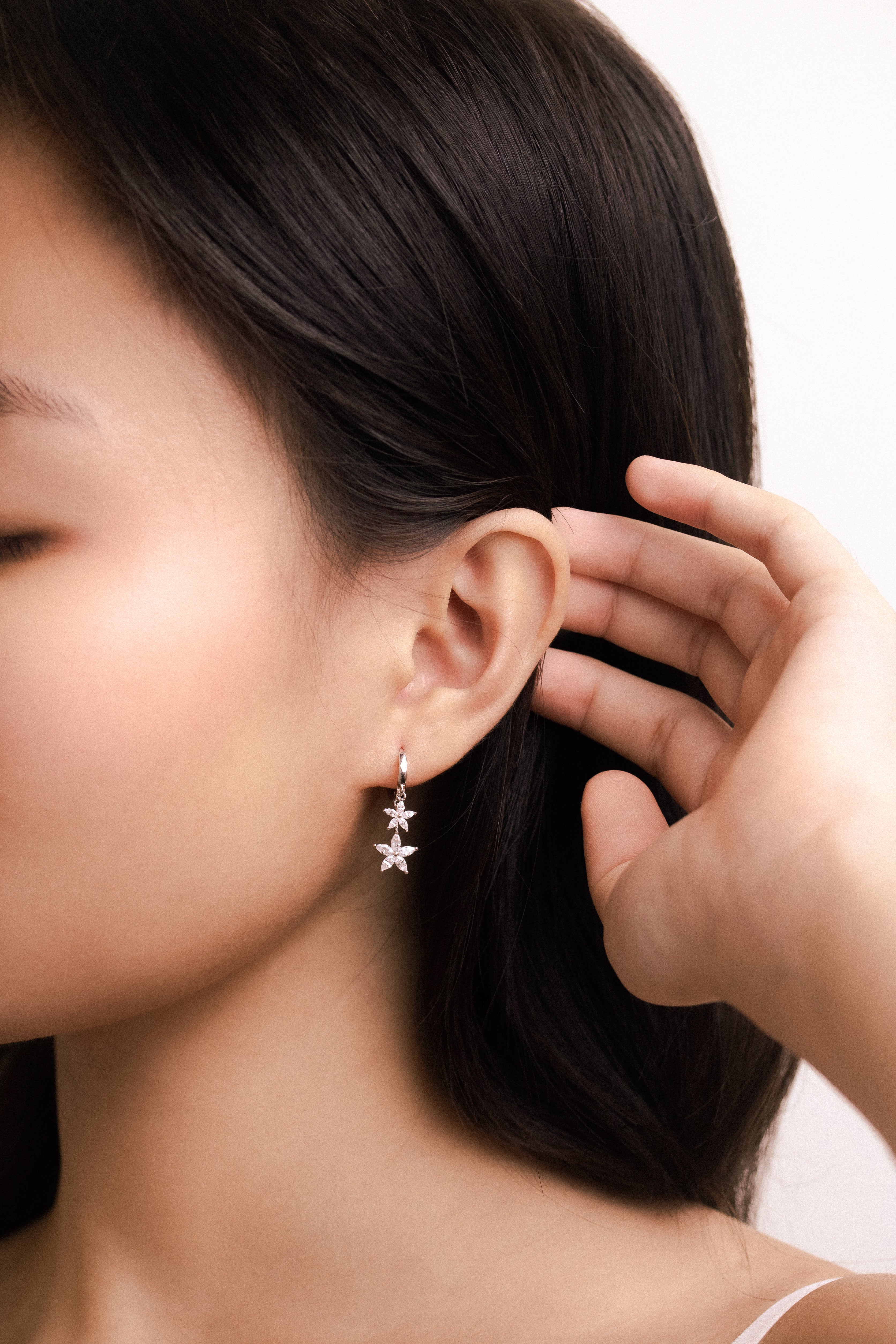 Fine jewellery, silver jewellery, hypoallergenic, tarnish-free,
Front full view of a silver flower-shaped adorned with crystal earrings worn on a person's ear with a hand adjusting it.