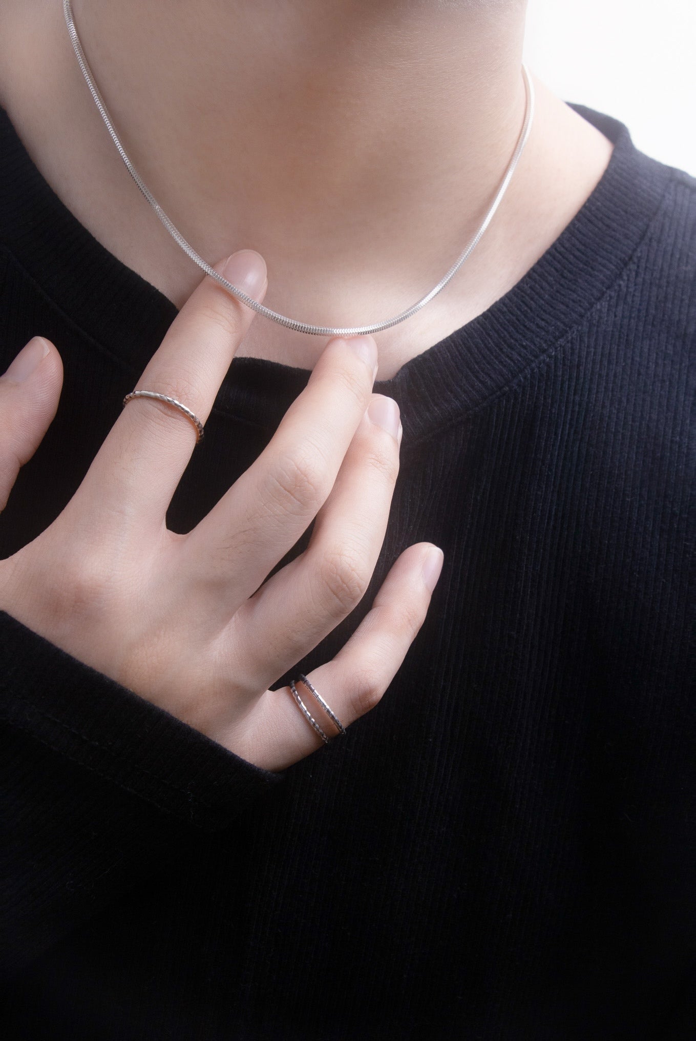 Person wearing a black shirt with a silver necklace and multiple silver rings on a plain black background, close up view