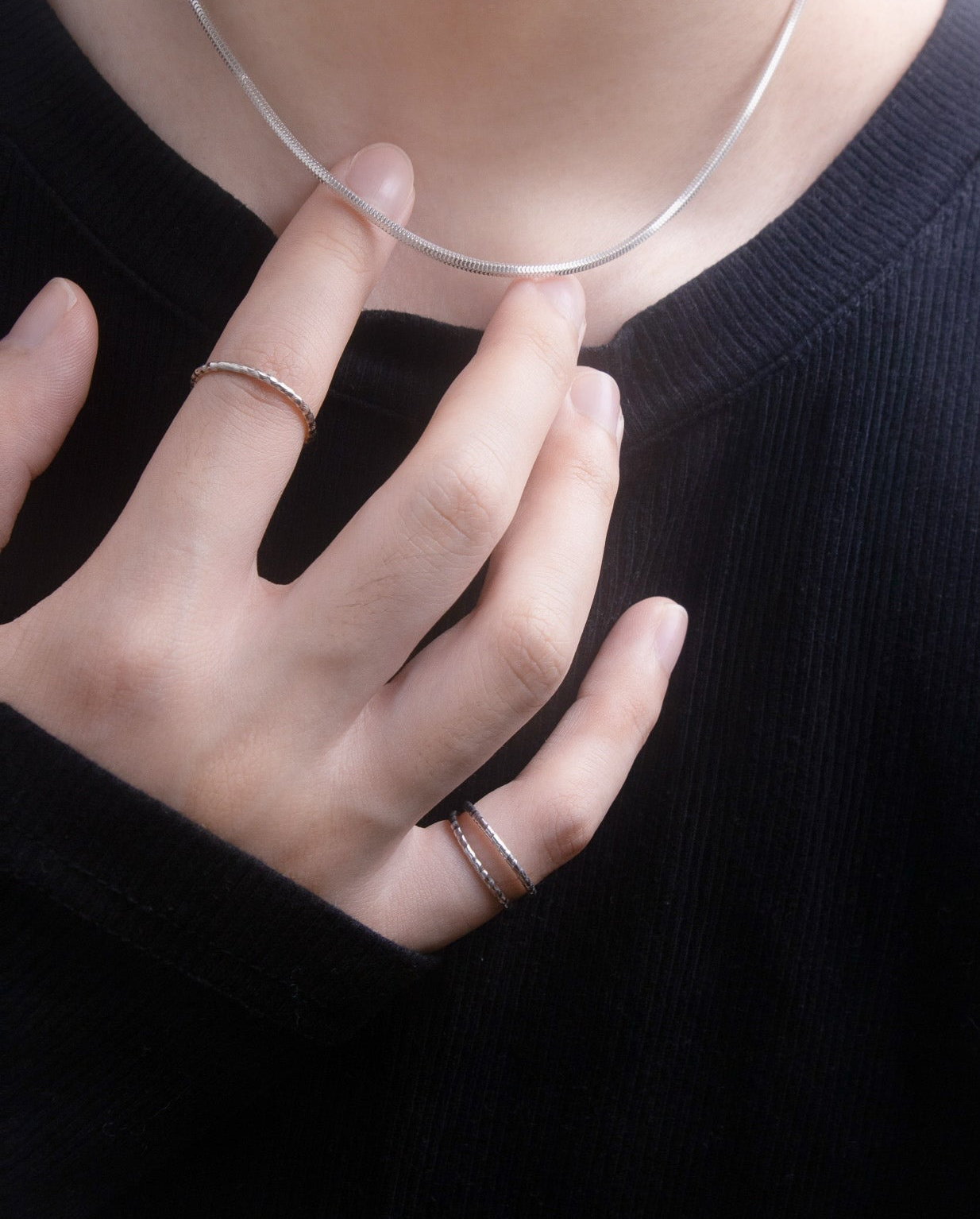 Person wearing a black shirt with a silver necklace and multiple silver rings on a plain black background, close up view