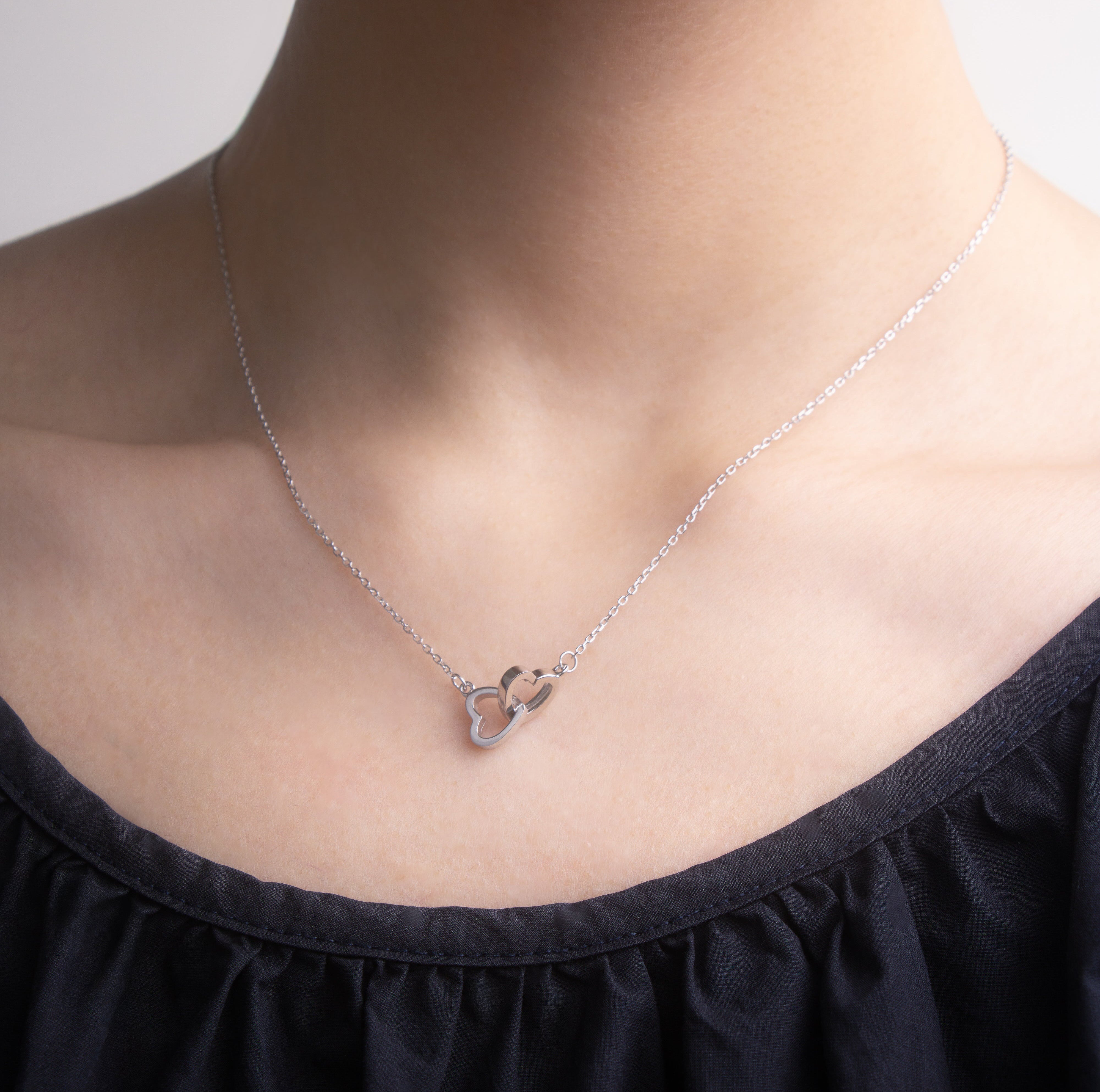 Close-up front view of a silver necklace with a heart-shaped pendant worn by a person in a black top.