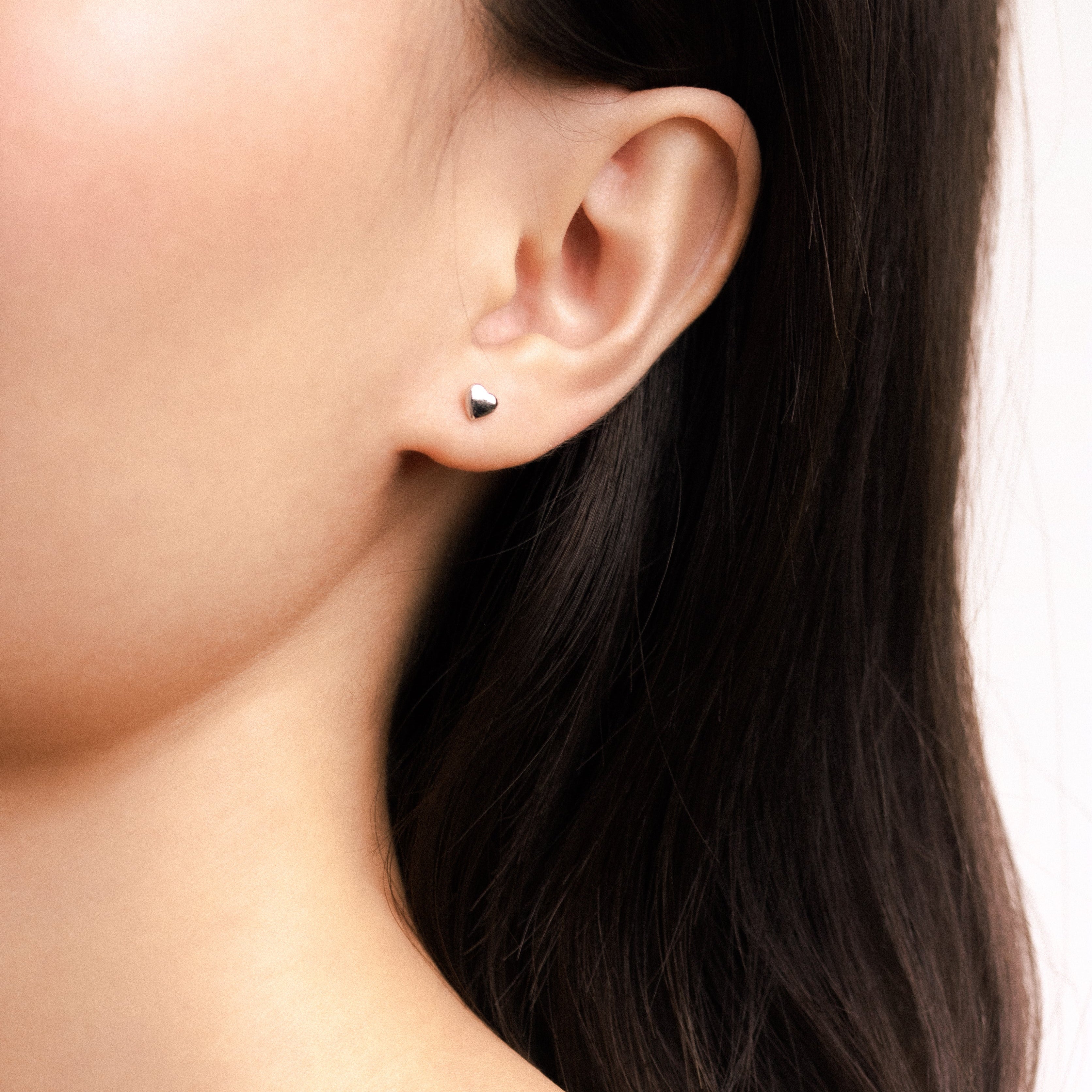 Fine jewellery, silver jewellery, hypoallergenic, tarnish-free, full view of a pair of Heart-shaped silver ear studs on a worn on a person's ear